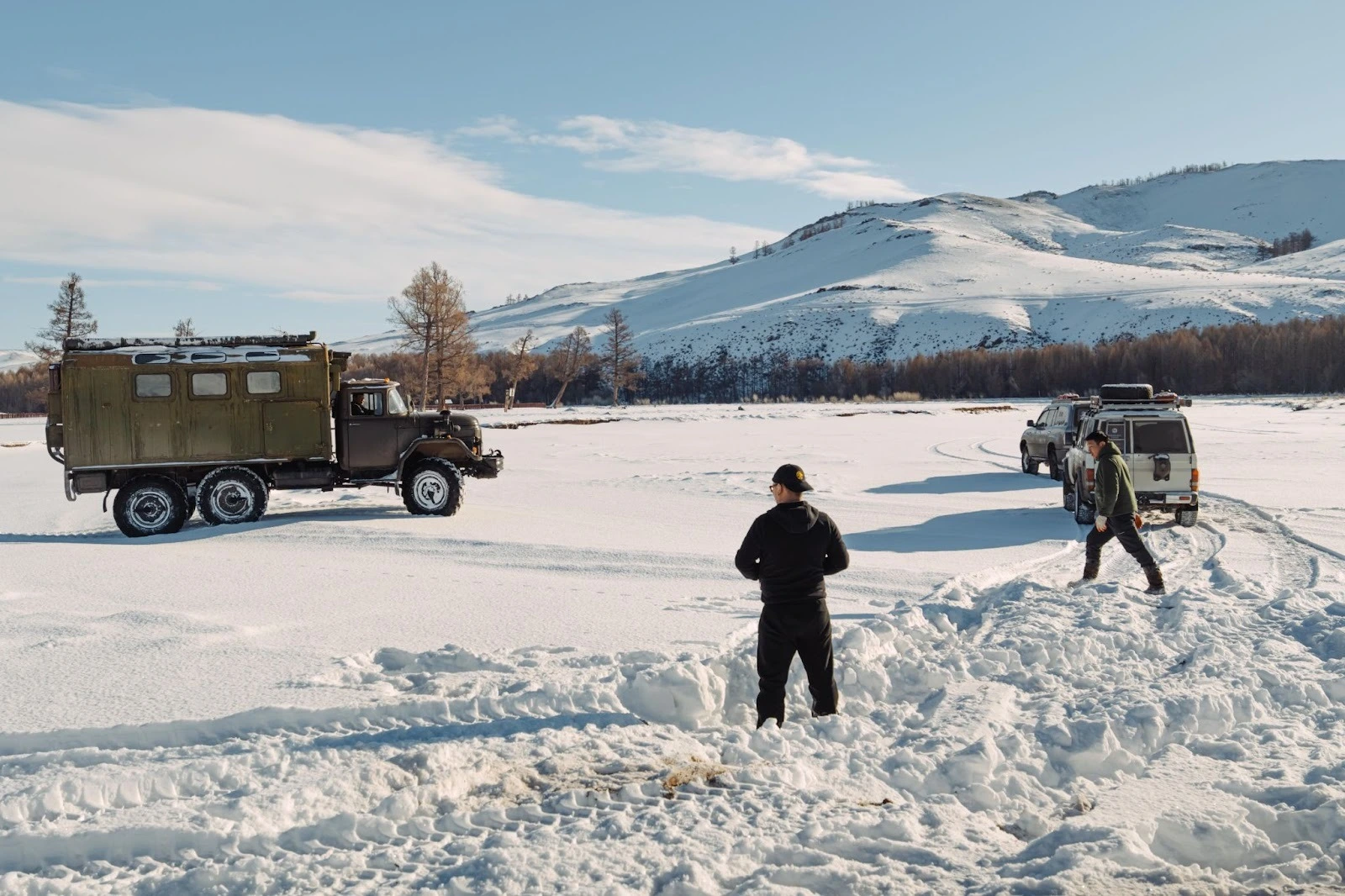 A large vintage green overland camper truck and two support off-road vehicles parked on a vast, snow-covered landscape with people standing nearby and snowy mountains in the background.
