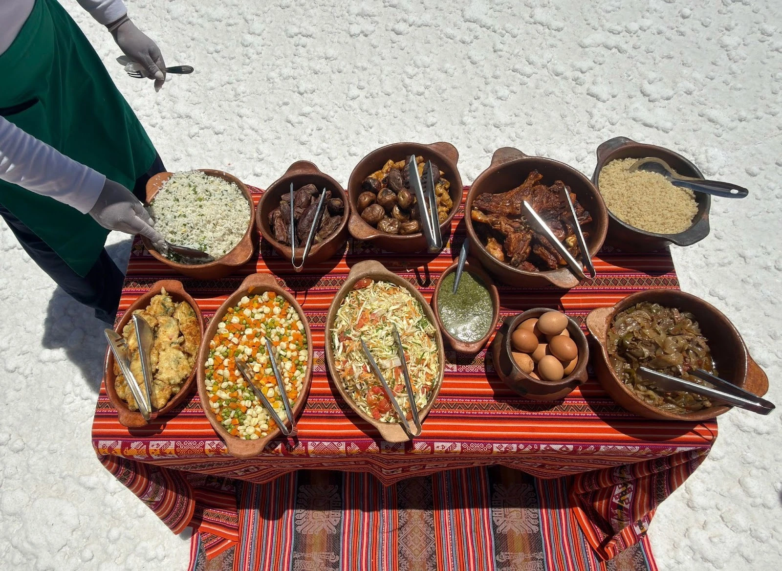 A traditional Andean lunch buffet served in clay pots on a colorful patterned textile table, set against the white landscape of the Salar de Uyuni salt flats.