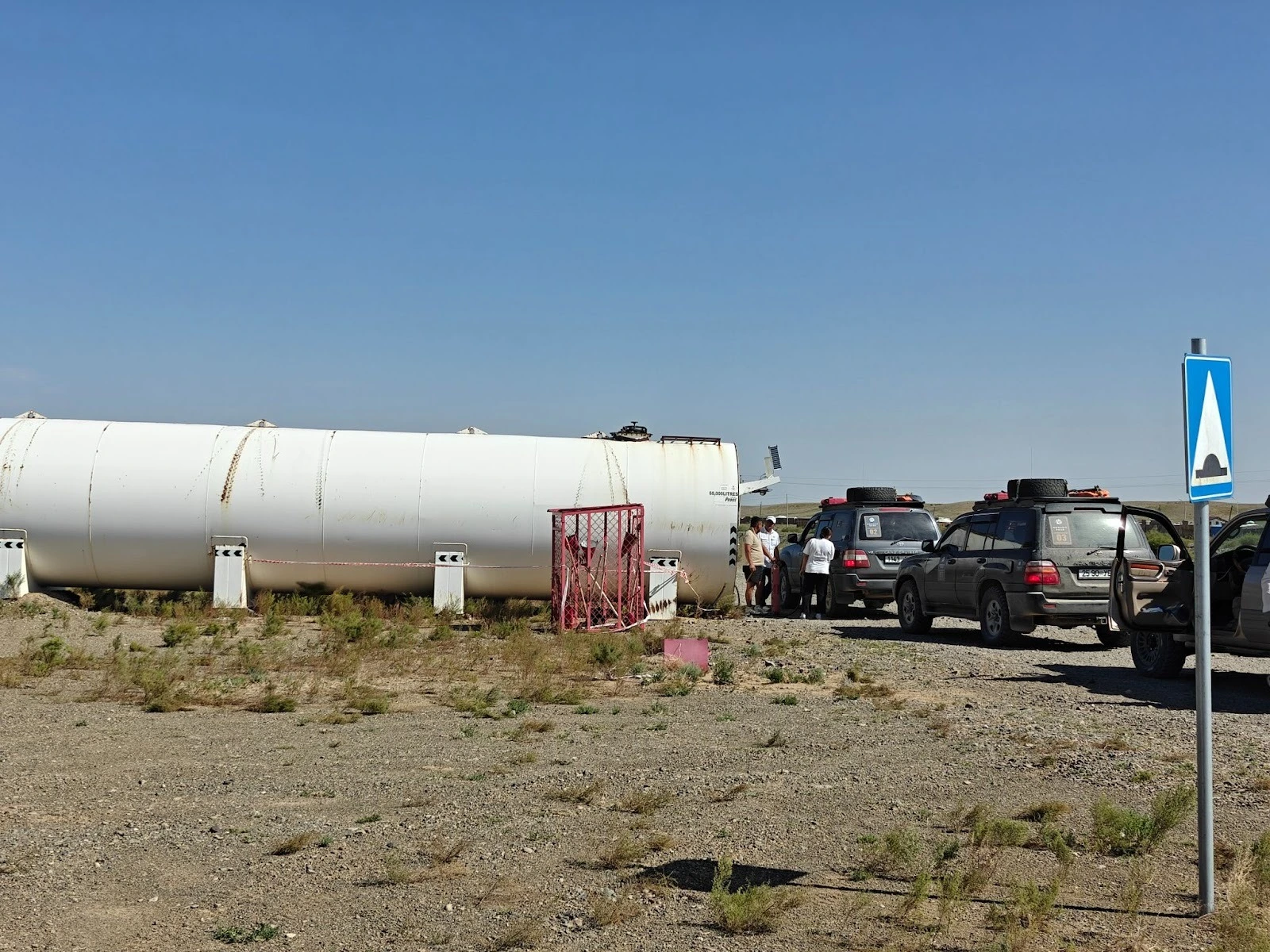 A large white cylindrical industrial storage tank lying in an arid desert field next to a group of off-road SUVs and travelers under a clear blue sky.
