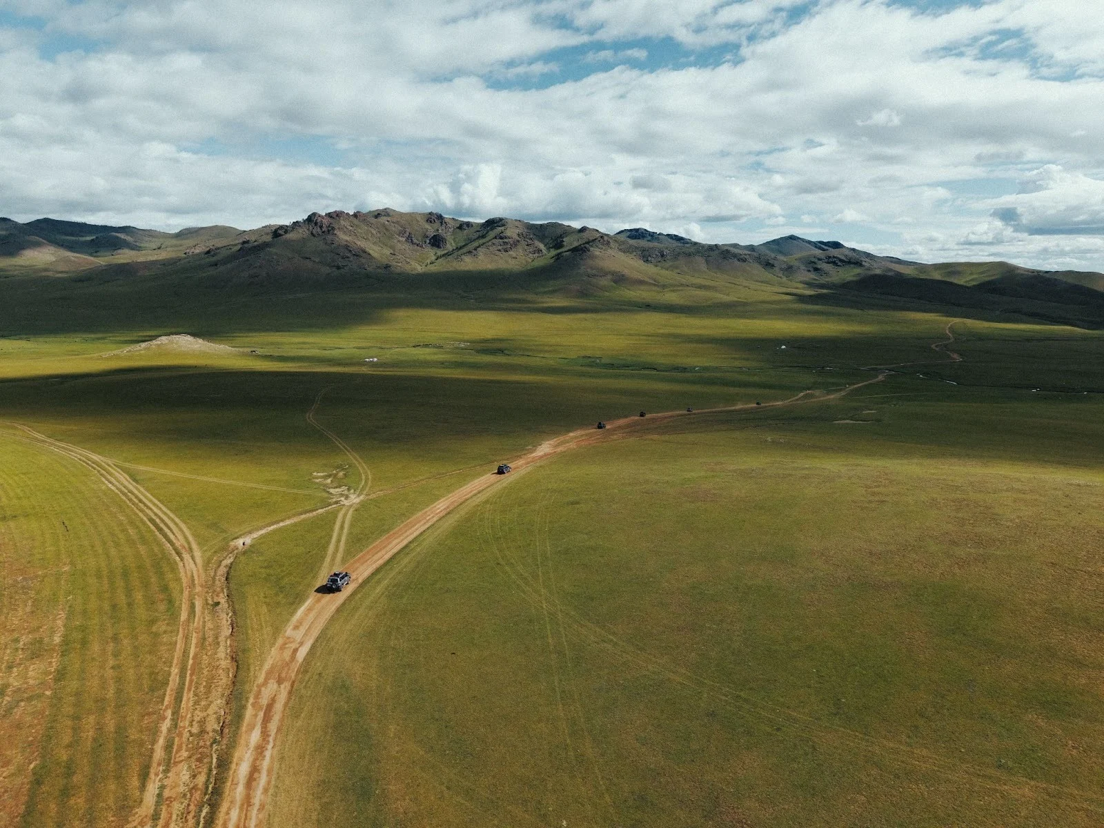 An aerial drone shot of a convoy of off-road vehicles driving along a winding dirt track through the vast, rolling green hills of the Mongolian steppe under a cloudy sky.