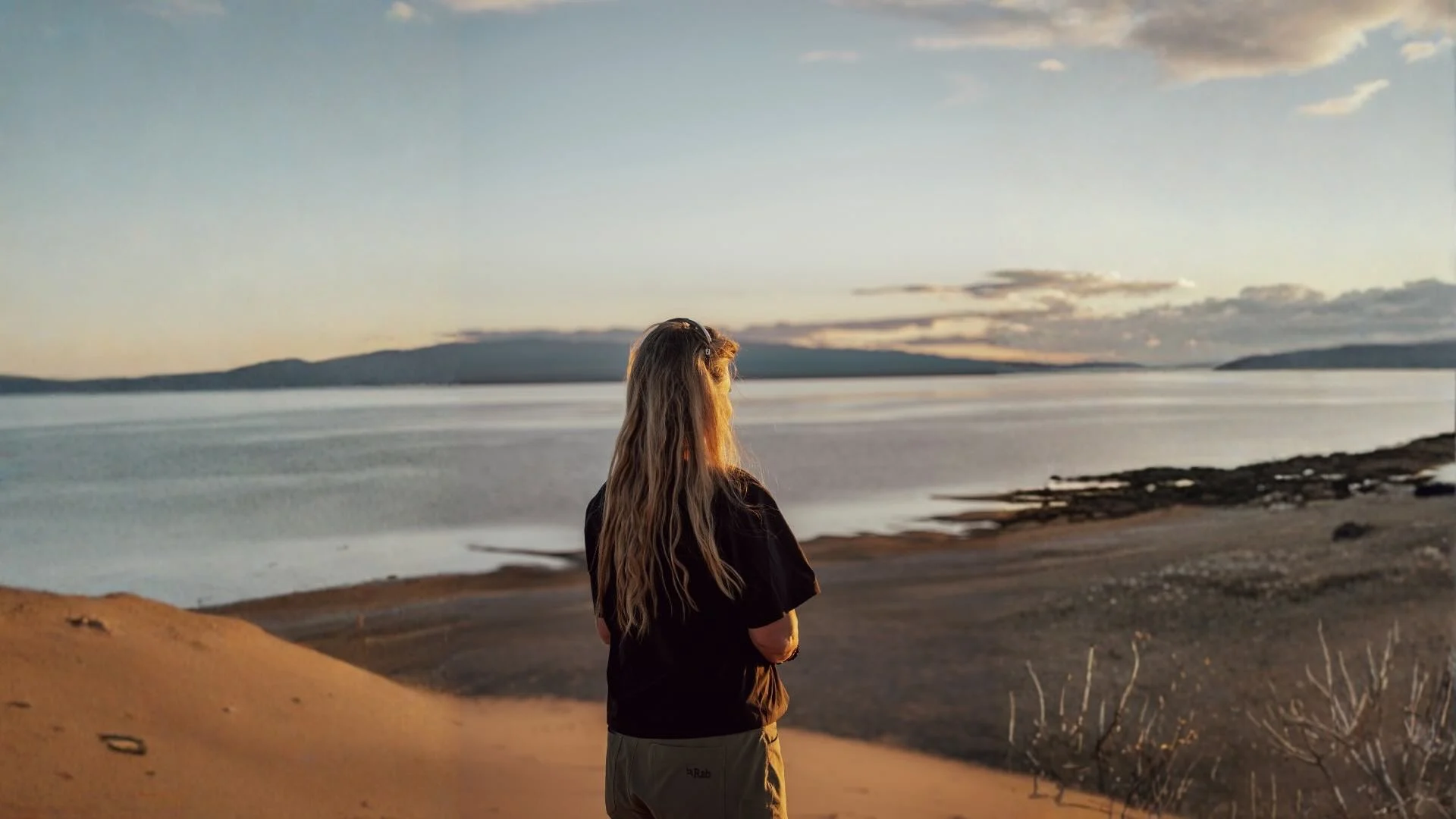 Back view of a female hiker in outdoor apparel standing on a sand dune, overlooking a vast coastline, rocky shores, and rolling hills under a soft sunset sky.