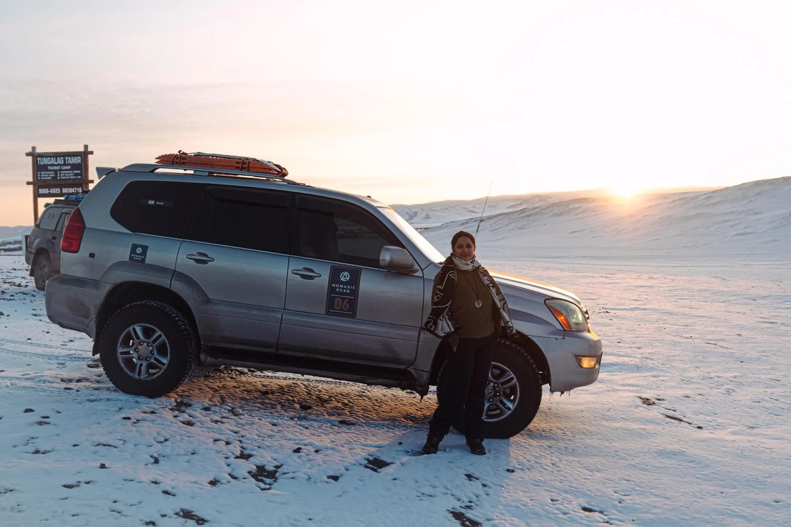 A silver off-road SUV with roof gear parked on a snow-covered terrain during sunset, next to a person standing by the passenger door and a wooden camp sign in the background.