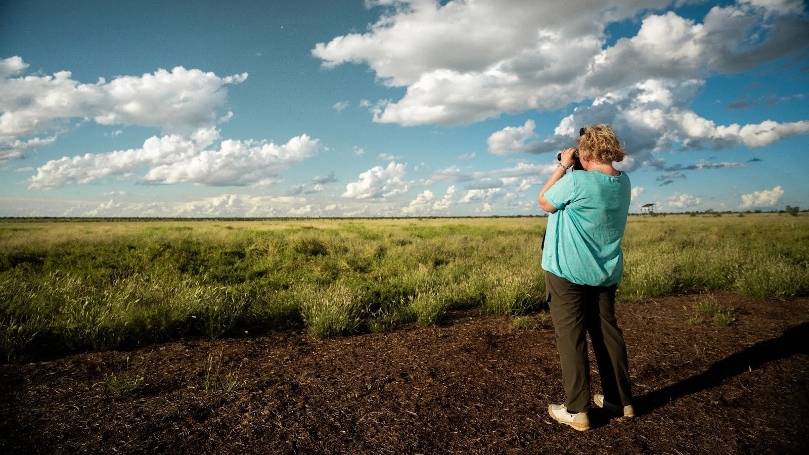 A woman stands in a grassy wetland field looking through binoculars under a cloudy blue sky.