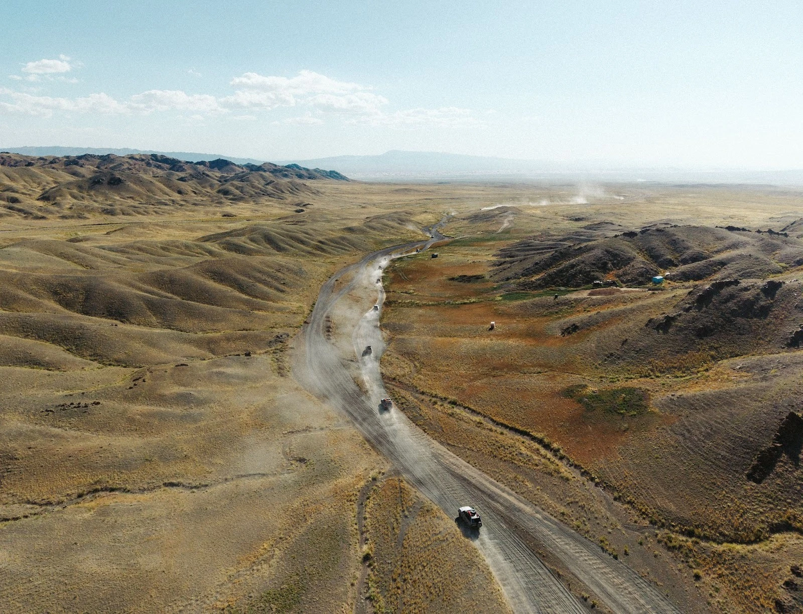 High-angle aerial drone view of a convoy of off-road 4x4 vehicles driving along a winding dirt road through the vast, arid, rolling hills of the Mongolian Gobi Desert under a clear sky.