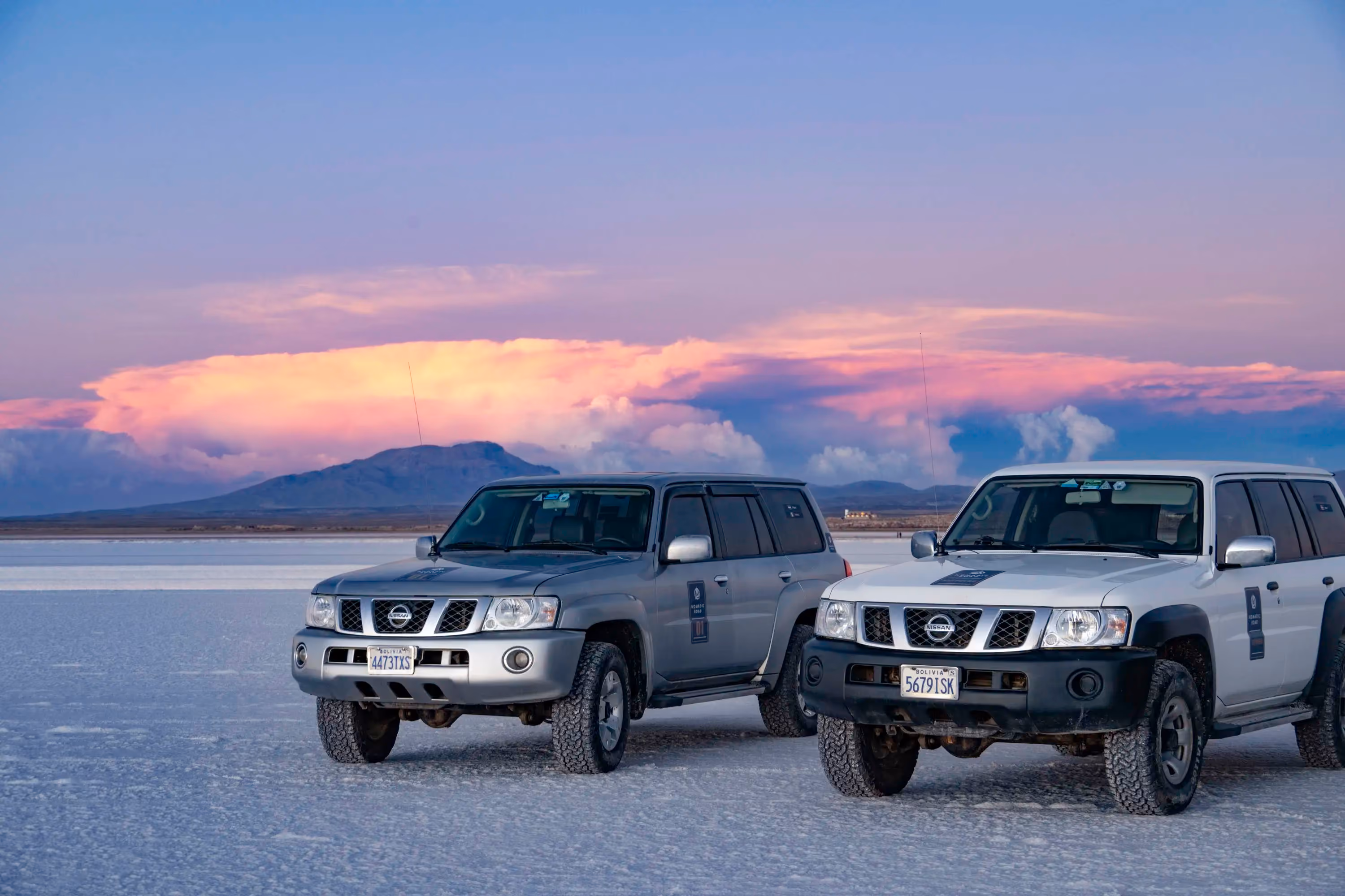 Tour SUVs parked on the Bolivian salt flats during a vibrant sunset with clouds over distant mountains.