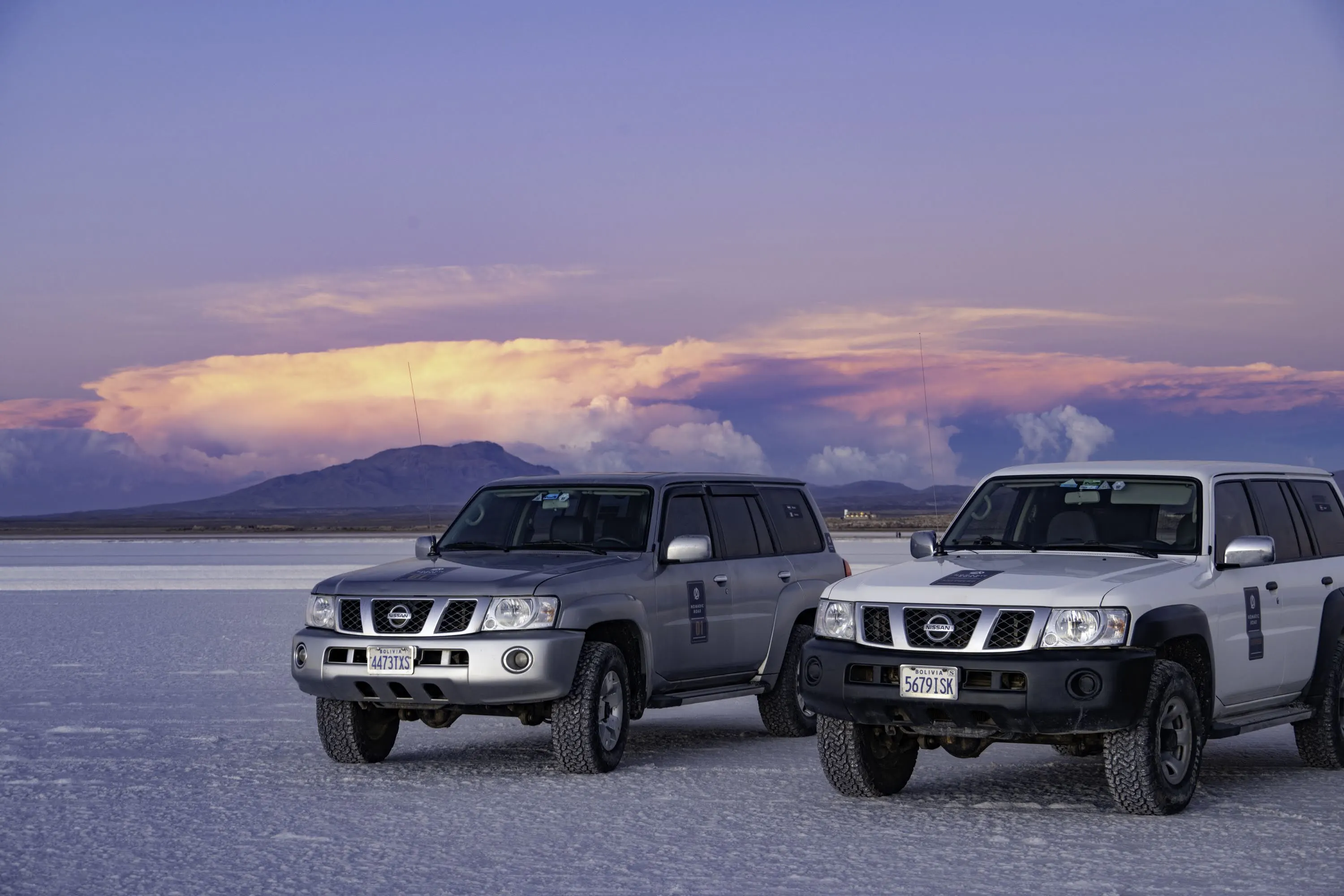 Tour SUVs parked on the Bolivian salt flats during a vibrant sunset with clouds over distant mountains.