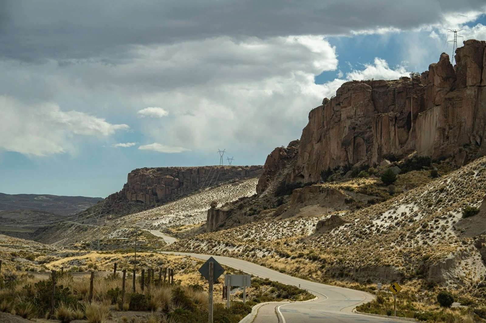A winding two-lane highway snakes through an arid, rocky canyon landscape in Bolivia under a cloudy sky with massive cliff formations and power lines on the ridges.