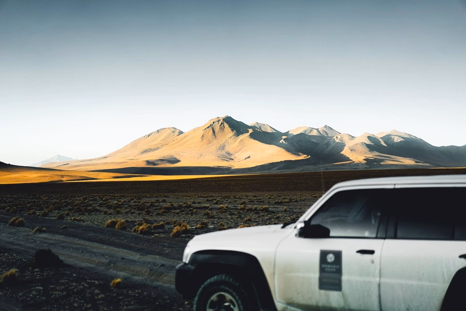 A white tour 4x4 vehicle driving along a remote dirt trail in the vast desert plains of the Bolivian Altiplano, with dramatic volcanic mountain peaks illuminated by warm sunlight in the background.