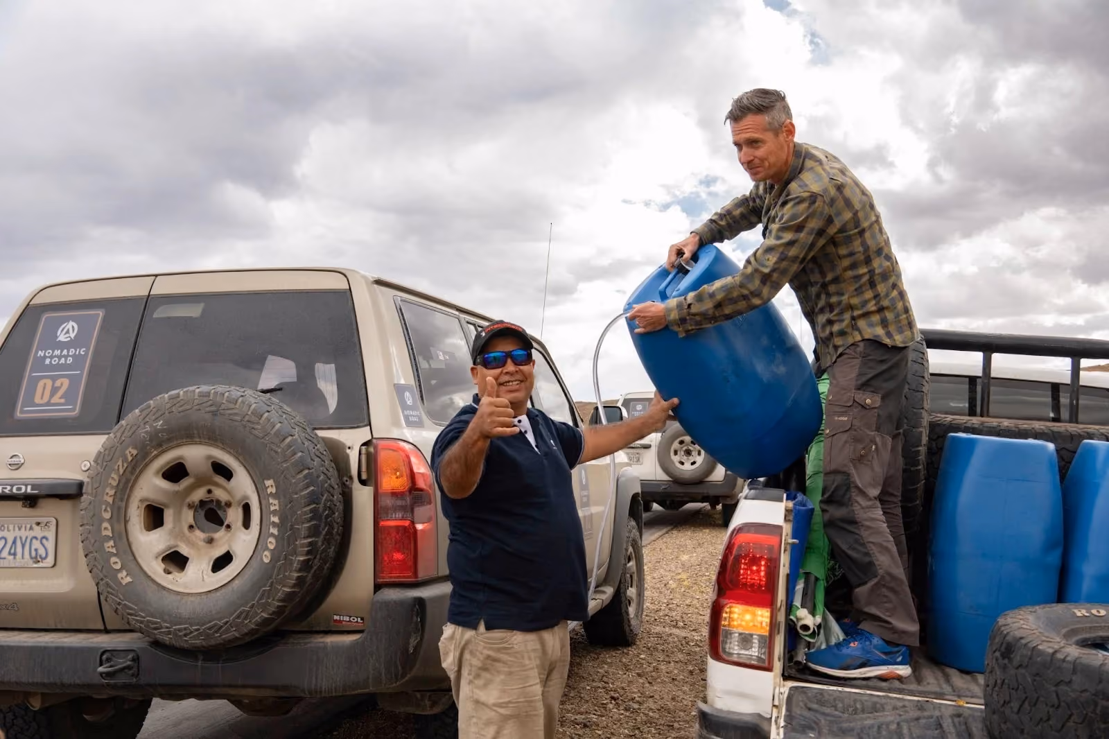Two men working together to siphon liquid from a large blue barrel in the bed of a pickup truck during a remote overland expedition under a cloudy sky.
