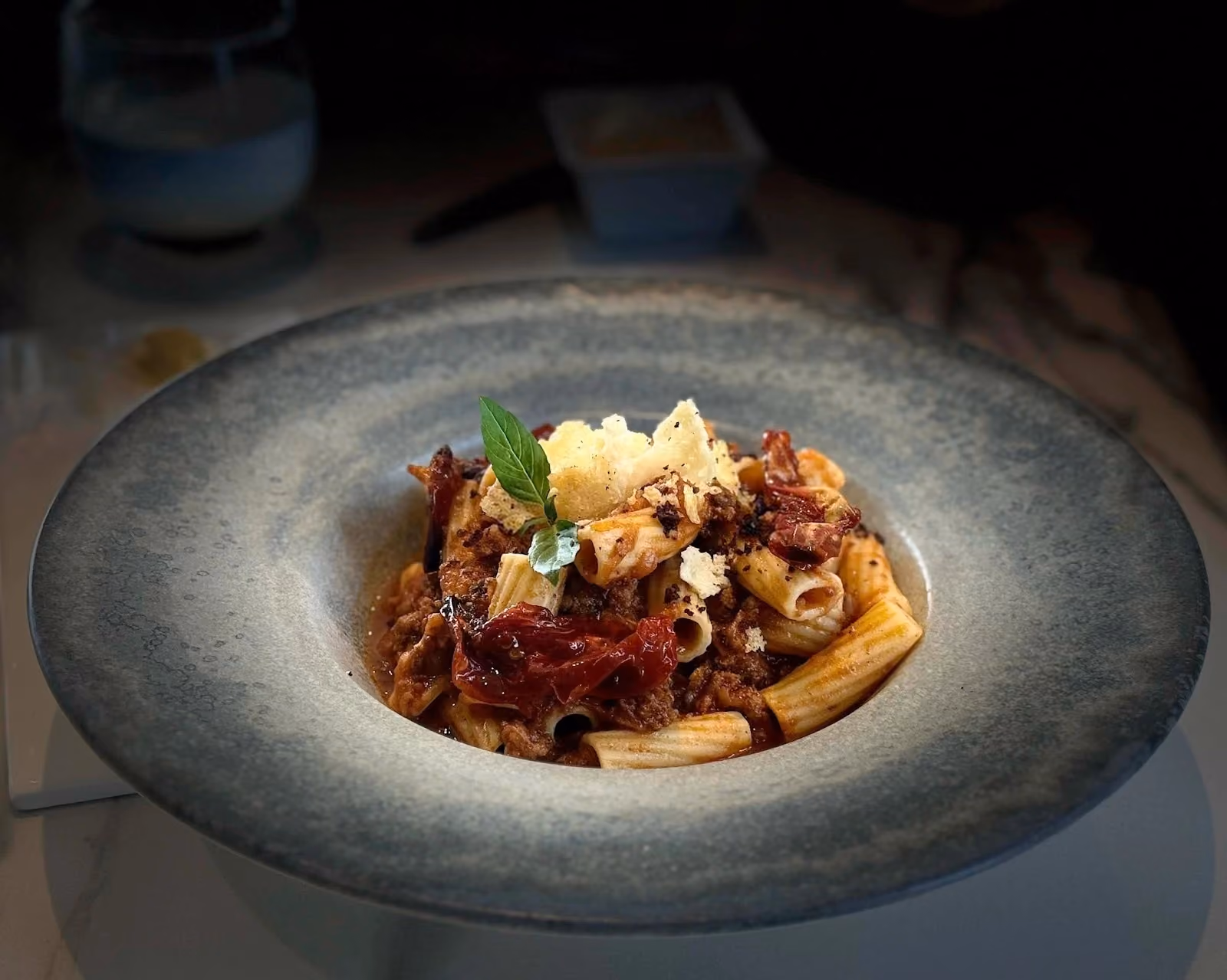 A gourmet dish of rigatoni pasta tossed in a rich meat ragù, served in a wide gray rimmed ceramic bowl and garnished with sun-dried tomatoes, grated cheese, and a fresh basil leaf.
