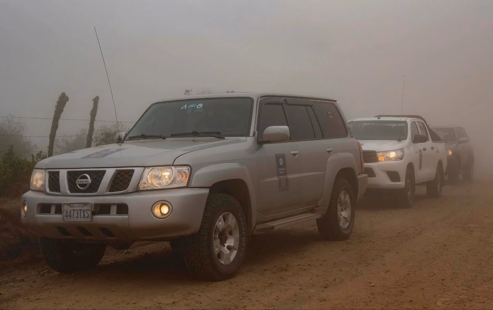 A silver 4x4 SUV with a Bolivian license plate leads a convoy of vehicles on a dirt road through thick fog.