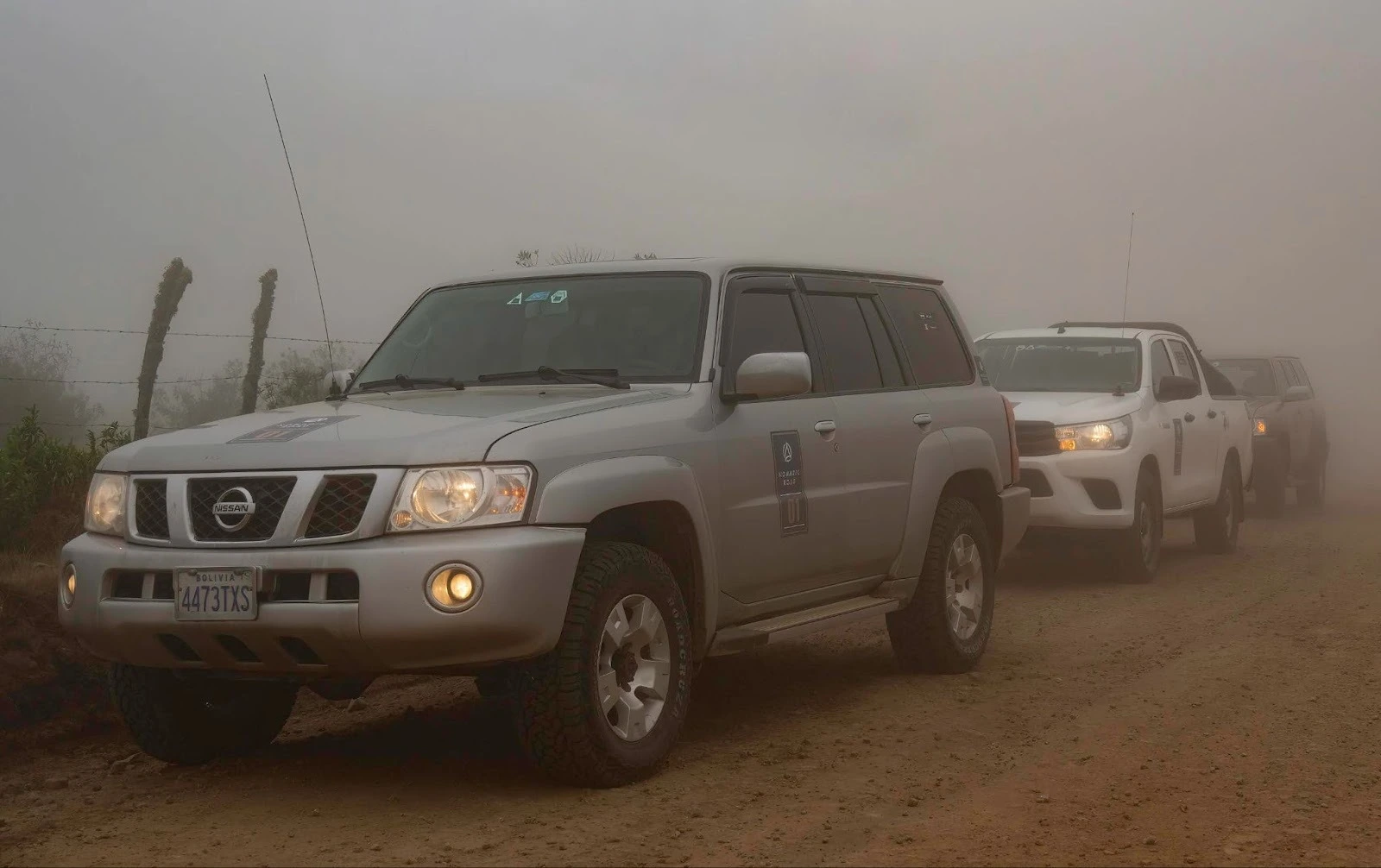 A silver 4x4 SUV with a Bolivian license plate leads a convoy of vehicles on a dirt road through thick fog.