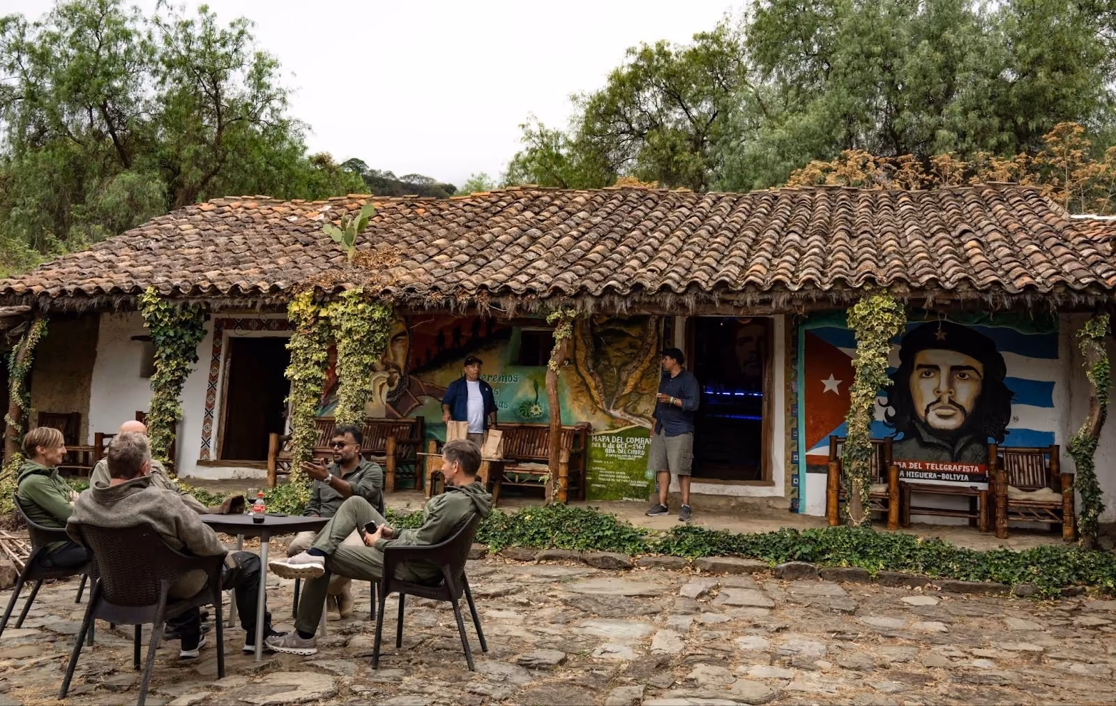 A historic stone building with a clay tile roof in La Higuera, Bolivia, featuring prominent murals of Che Guevara and outdoor seating where visitors are resting.