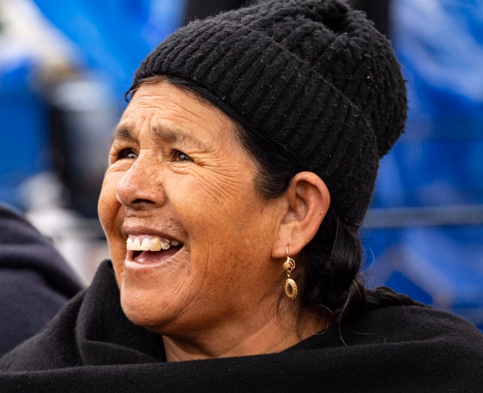 Close-up portrait of a smiling woman looking upward, wearing a black ribbed-knit beanie, gold filigree dangle earrings, and a dark black shawl against a blurred blue background.