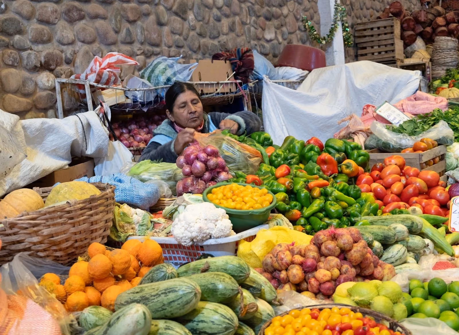 A local woman vendor sits behind a vibrant and dense display of fresh agricultural produce, including potatoes, peppers, onions, and cauliflower at a traditional open-air market with a stone wall background.