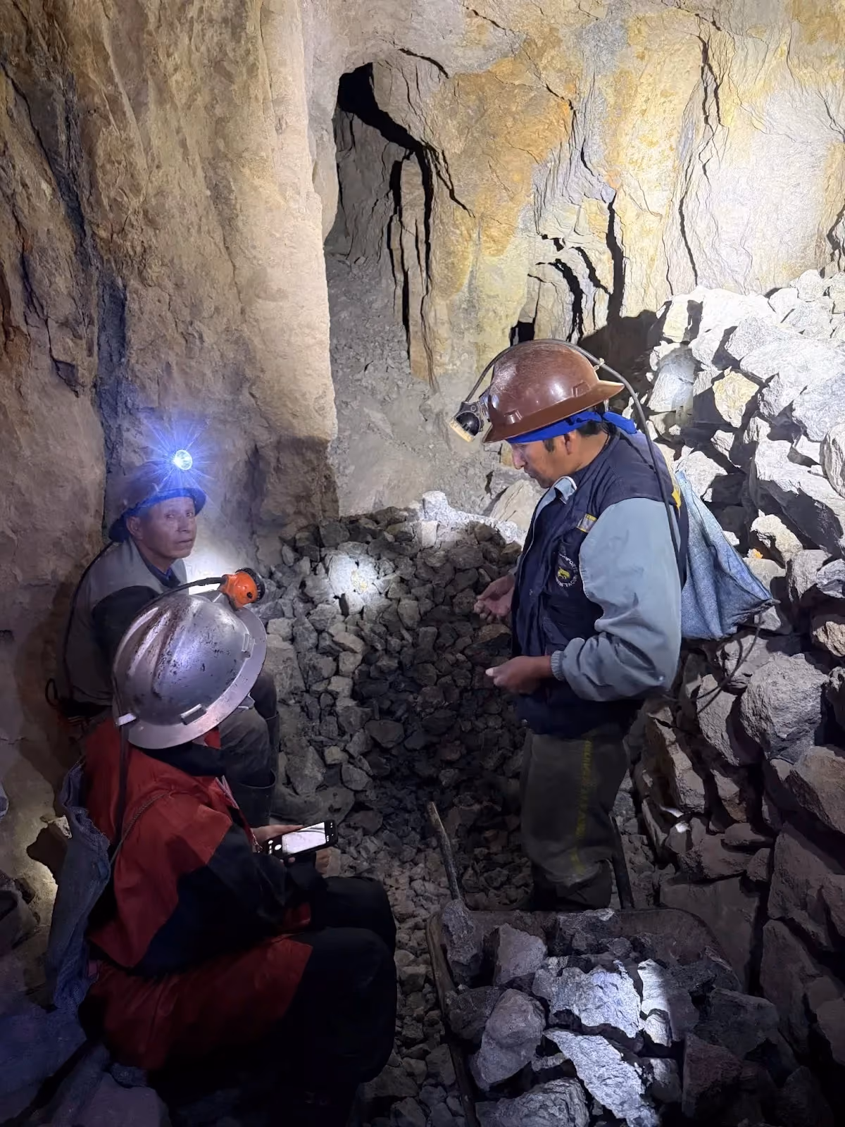 Three miners wearing hard hats with headlamps talk inside a narrow, rocky underground tunnel surrounded by stone walls and loose debris.