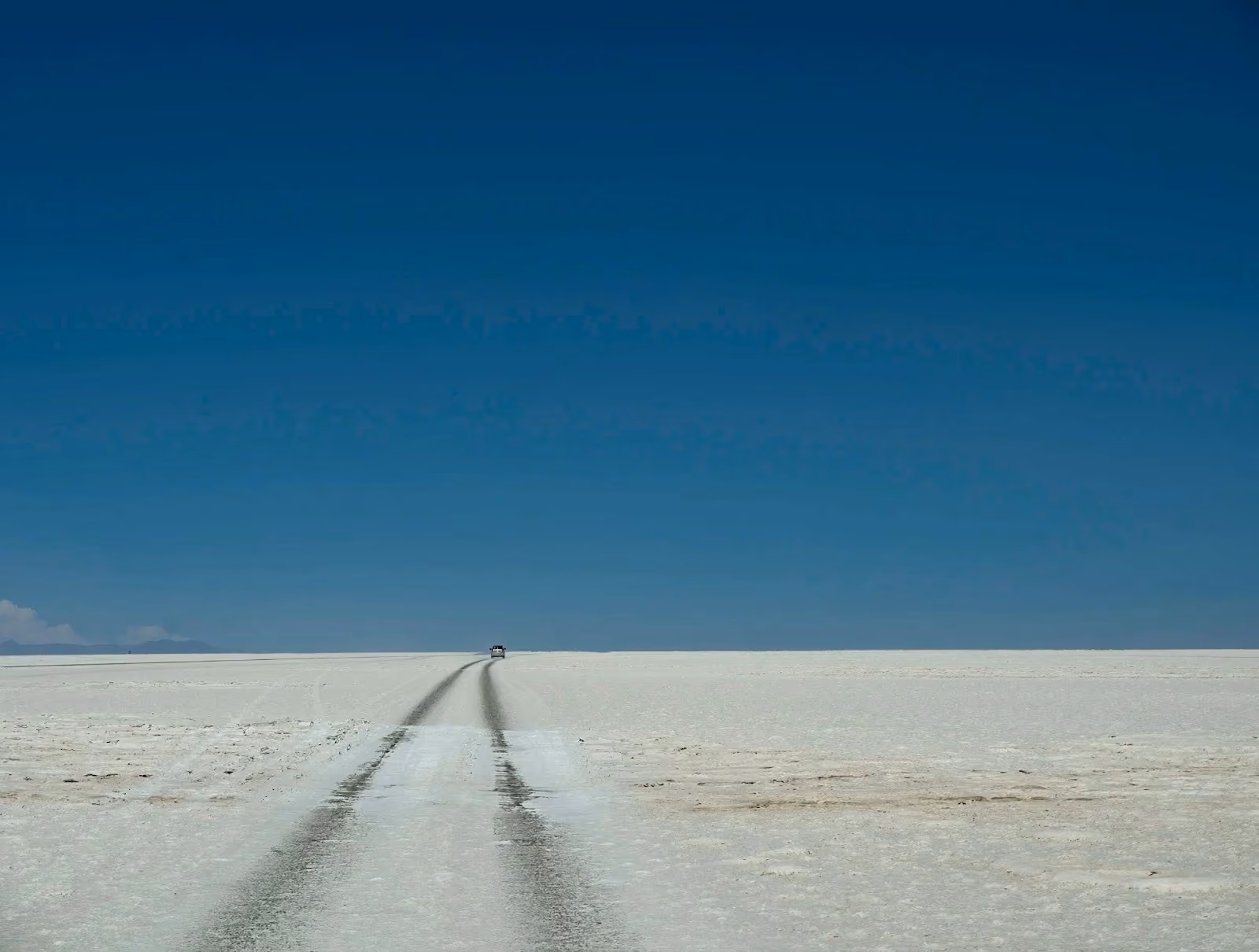 A wide landscape view of the vast, white Salar de Uyuni salt flats in Bolivia under a clear blue sky, featuring dark vehicle tracks stretching toward the distant horizon.