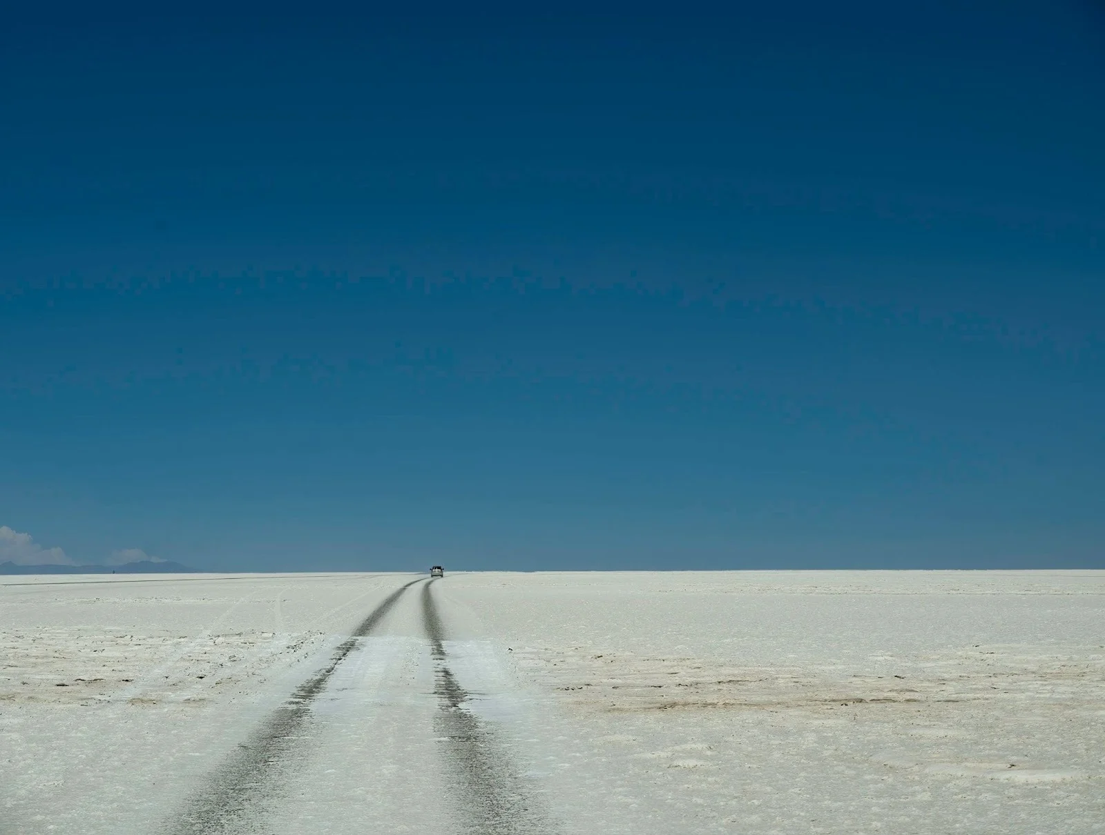 A wide landscape view of the vast, white Salar de Uyuni salt flats in Bolivia under a clear blue sky, featuring dark vehicle tracks stretching toward the distant horizon.