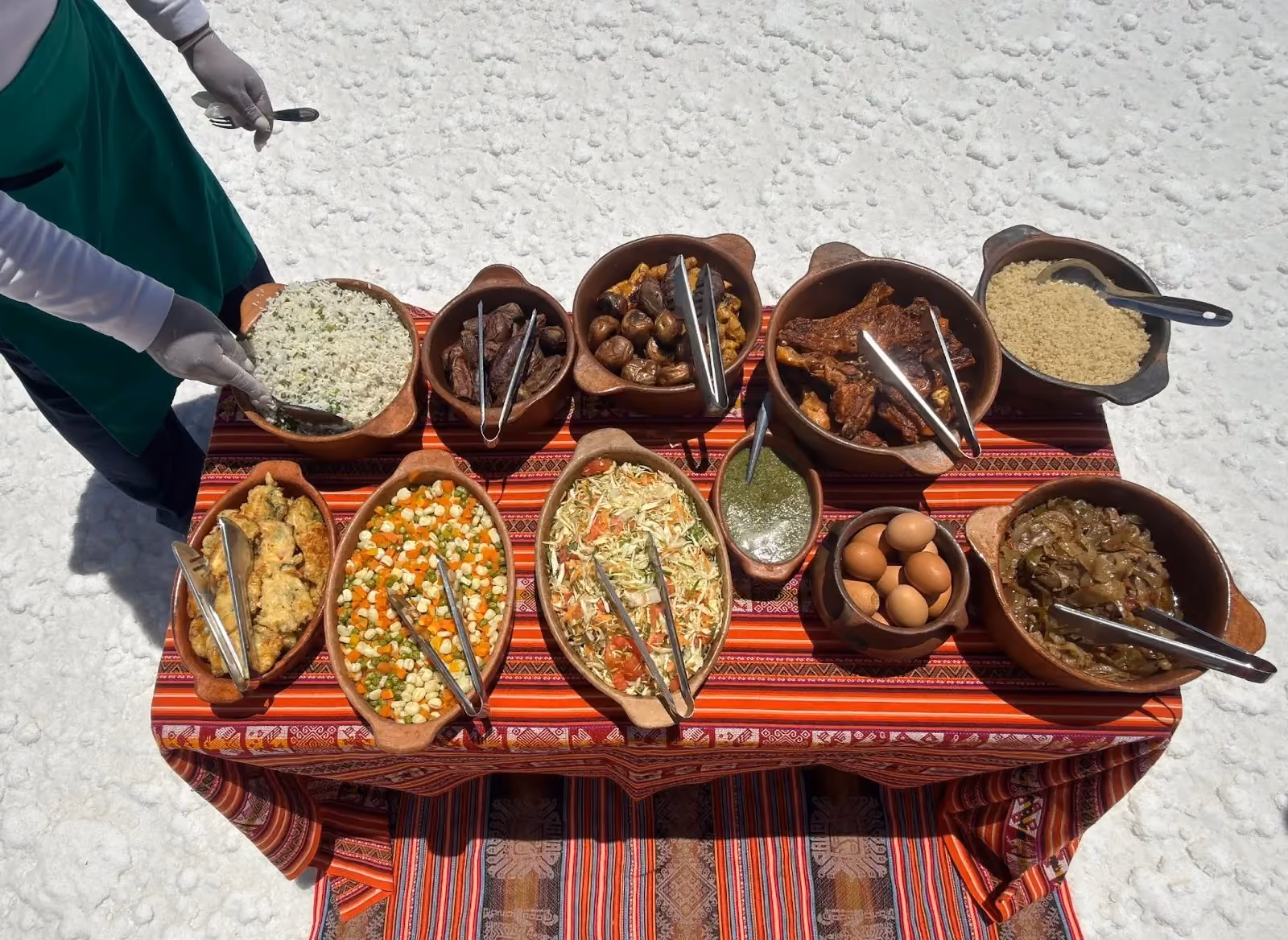 A gourmet buffet lunch spread of traditional Bolivian food in clay bowls arranged on a colorful Andean textile table in the middle of a bright white salt desert.