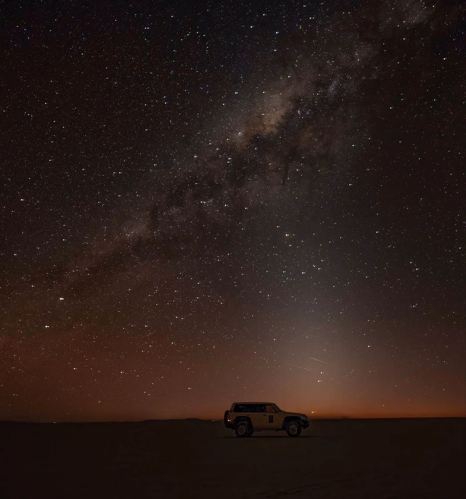 The Milky Way galaxy band intersects with a bright, vertical cone of zodiacal light in a star-filled night sky over a dark desert horizon with a vehicle silhouette.