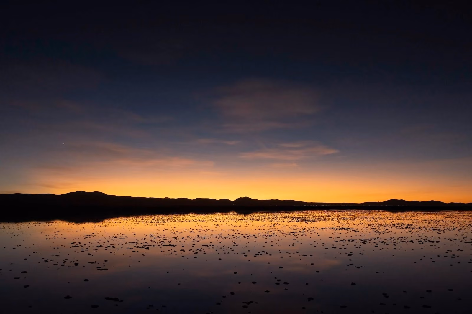A vast, flooded salt flat reflecting a vibrant orange and dark blue sunset sky over low mountain ridges in the distance.
