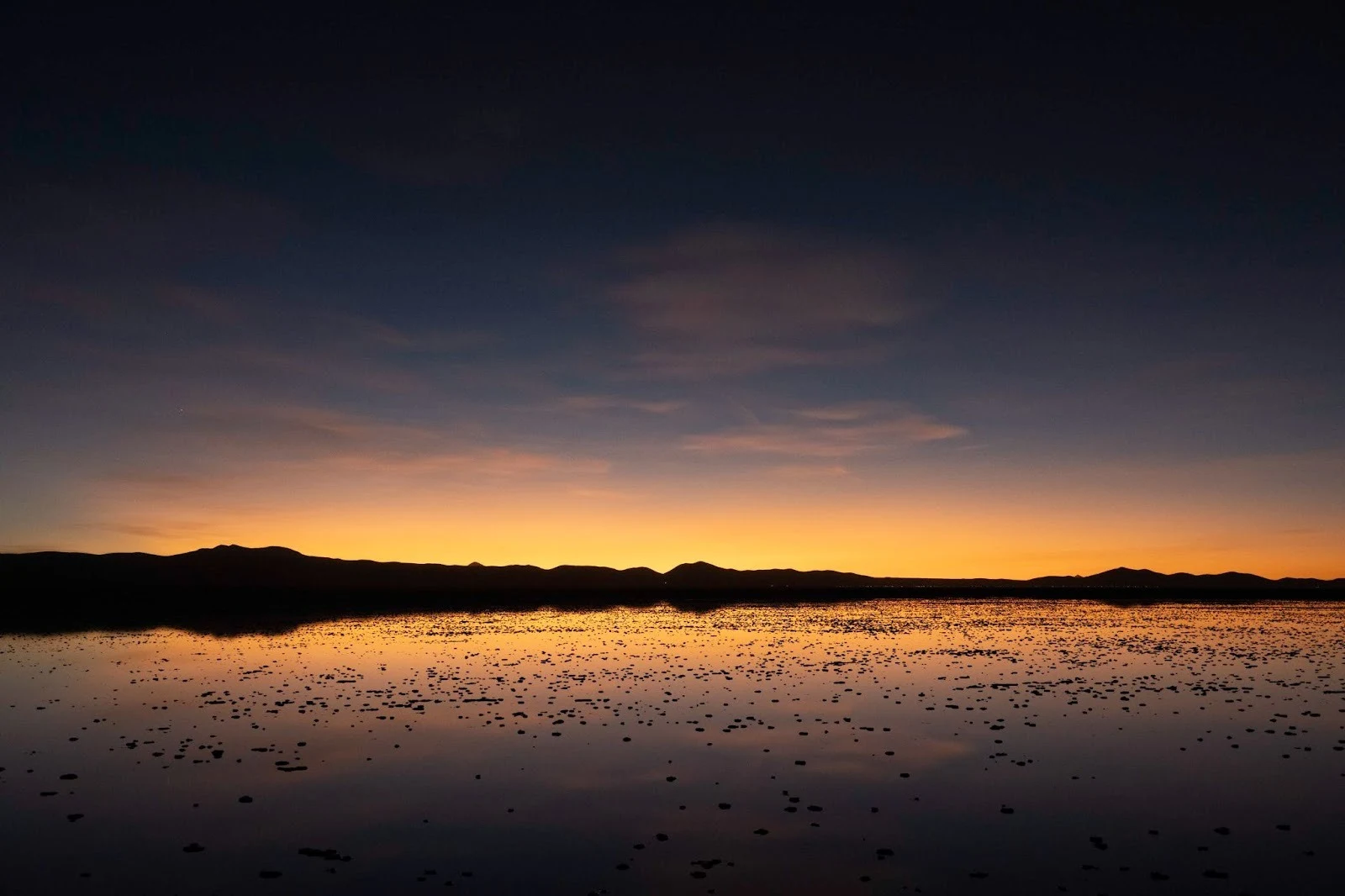 A vast, flooded salt flat reflecting a vibrant orange and dark blue sunset sky over low mountain ridges in the distance.