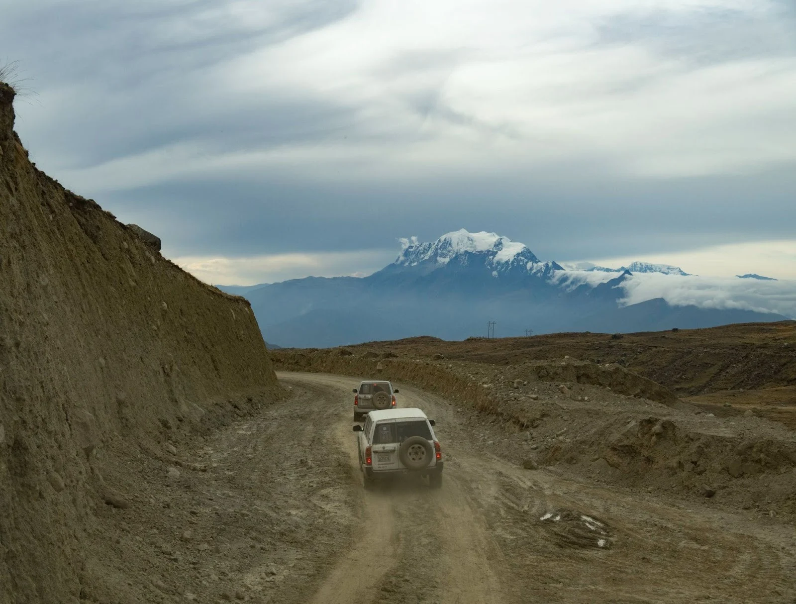Two 4WD vehicles traveling along a dusty, unpaved mountain road on a barren plateau with a snow-capped Himalayan mountain peak under a cloudy sky in Upper Mustang, Nepal.