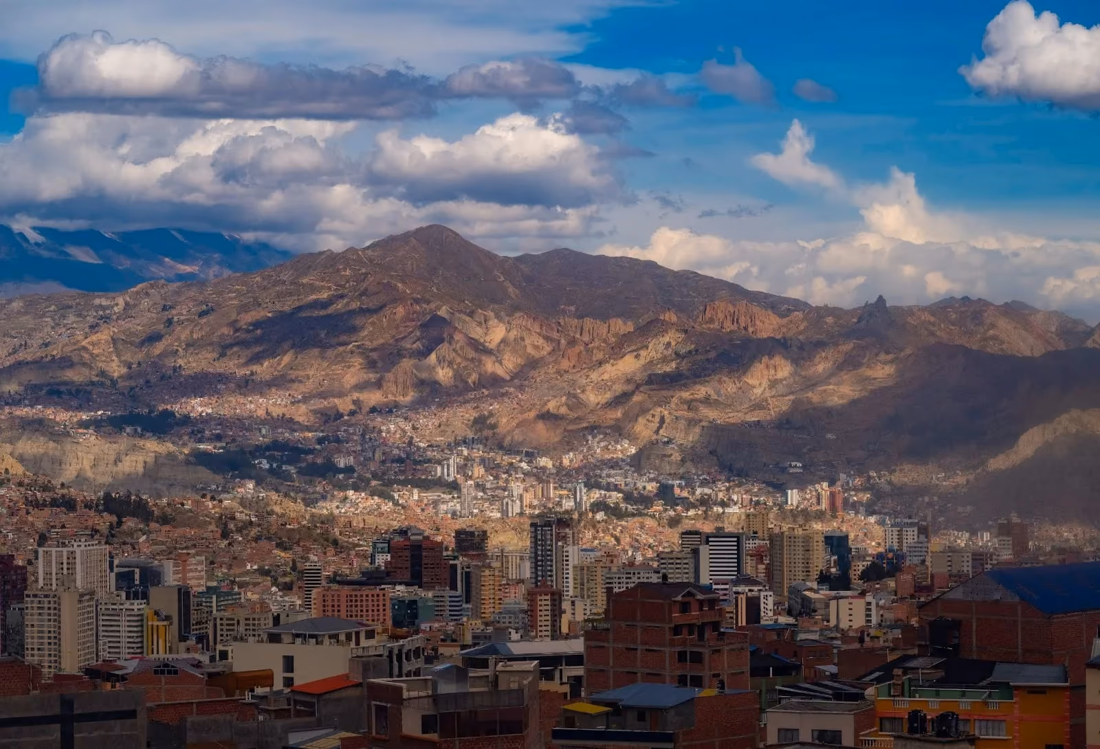 Panoramic view of the high-altitude city of La Paz, Bolivia, densely packed into a canyon valley with steep, rugged mountain slopes and white clouds in the blue sky above.