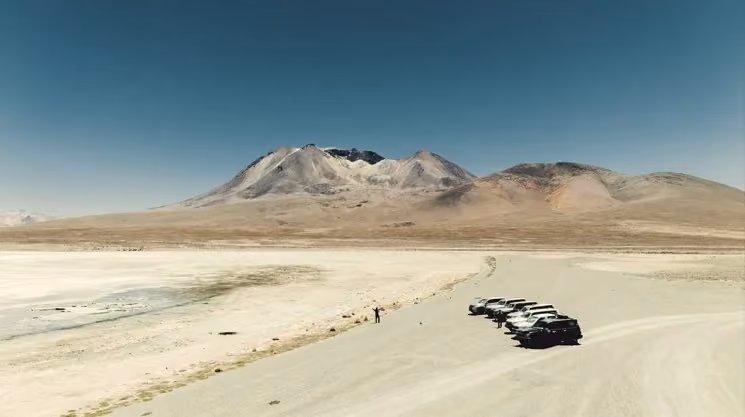 A caravan of dark overland vehicles traversing an arid, gravelly plain toward a snow-dusted volcano under a clear blue sky in the high-altitude desert.