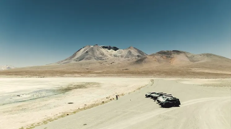 A caravan of dark overland vehicles traversing an arid, gravelly plain toward a snow-dusted volcano under a clear blue sky in the high-altitude desert.
