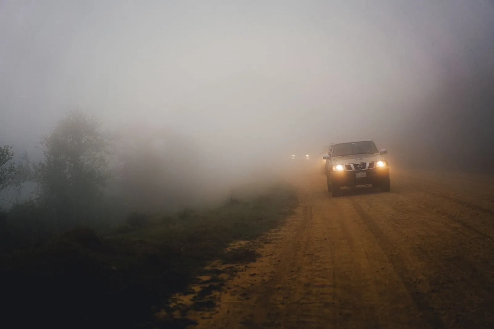 Head-on view of a car driving on an unpaved dirt road enveloped in thick, heavy fog with its headlights turned on to illuminate the misty path ahead.