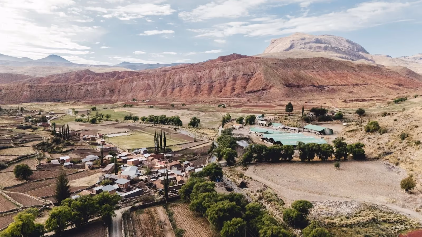 High-angle drone photograph showing a small Andean village with adobe houses, green farm plots, and massive reddish-brown layered mountains under a partly cloudy sky.