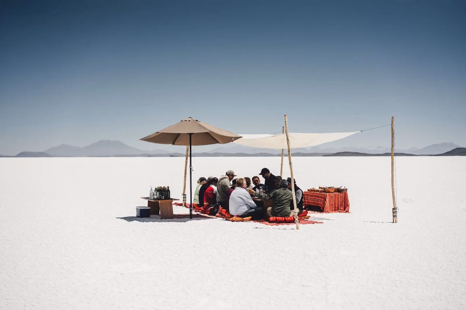 A group of people sitting on red cushions around a low picnic table under a sun umbrella and fabric awning, enjoying an outdoor gourmet meal in the middle of a vast white desert under a clear blue sky.