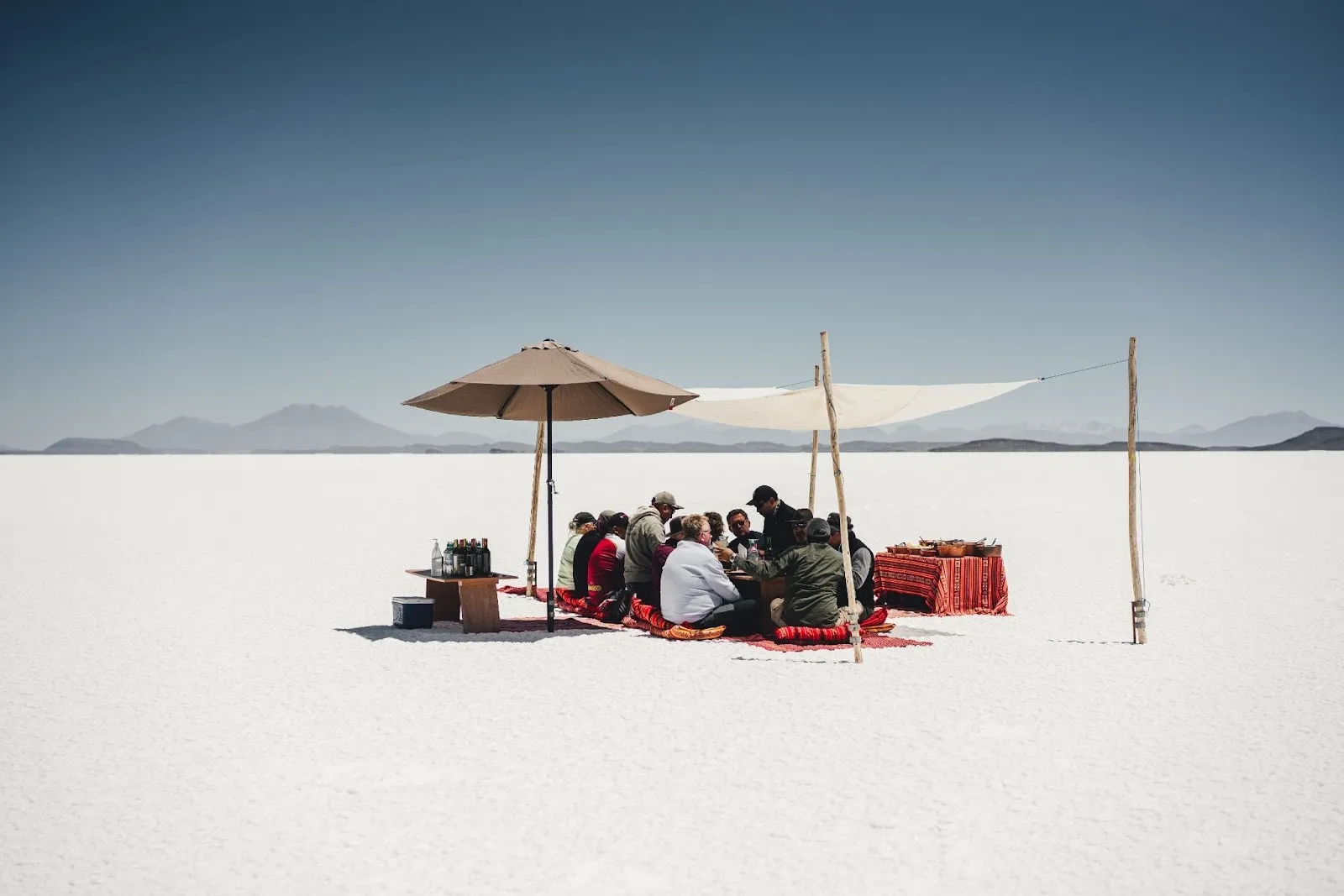 A group of people sitting on red cushions around a low picnic table under a sun umbrella and fabric awning, enjoying an outdoor gourmet meal in the middle of a vast white desert under a clear blue sky.