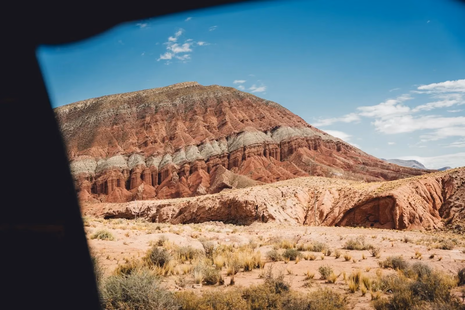 A striped red and white rock mountain rises behind an arid desert plain with scattered shrubs under a clear blue sky.