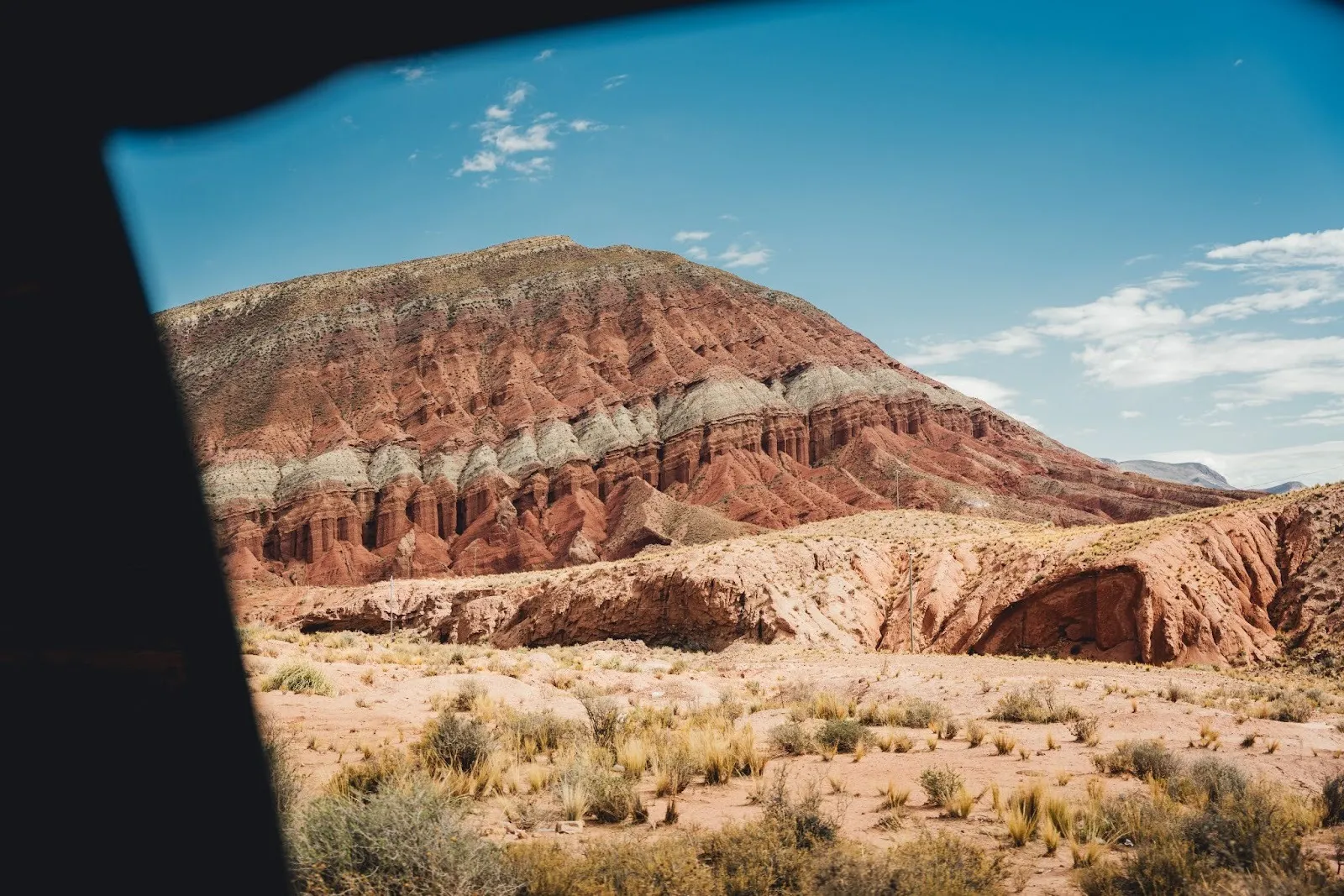 A striped red and white rock mountain rises behind an arid desert plain with scattered shrubs under a clear blue sky.