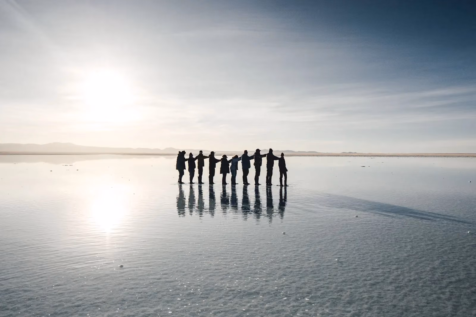 Group of people standing hand-in-hand reflecting on the flooded Salar de Uyuni salt flat.