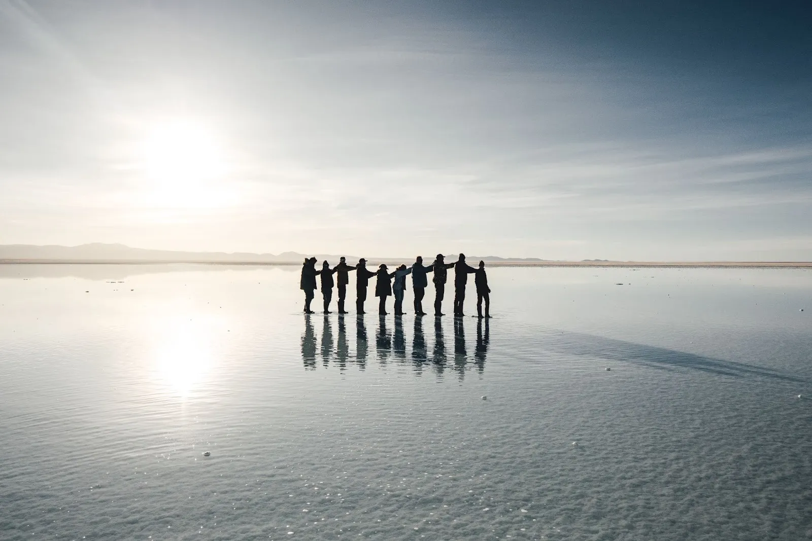 Group of people standing hand-in-hand reflecting on the flooded Salar de Uyuni salt flat.