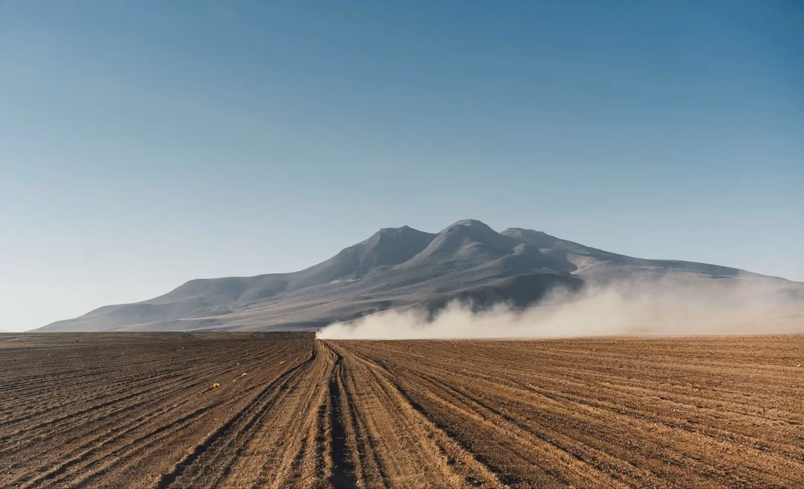 Dusty vehicle trail on a corrugated dirt road leading toward a volcanic mountain range under a clear sky.