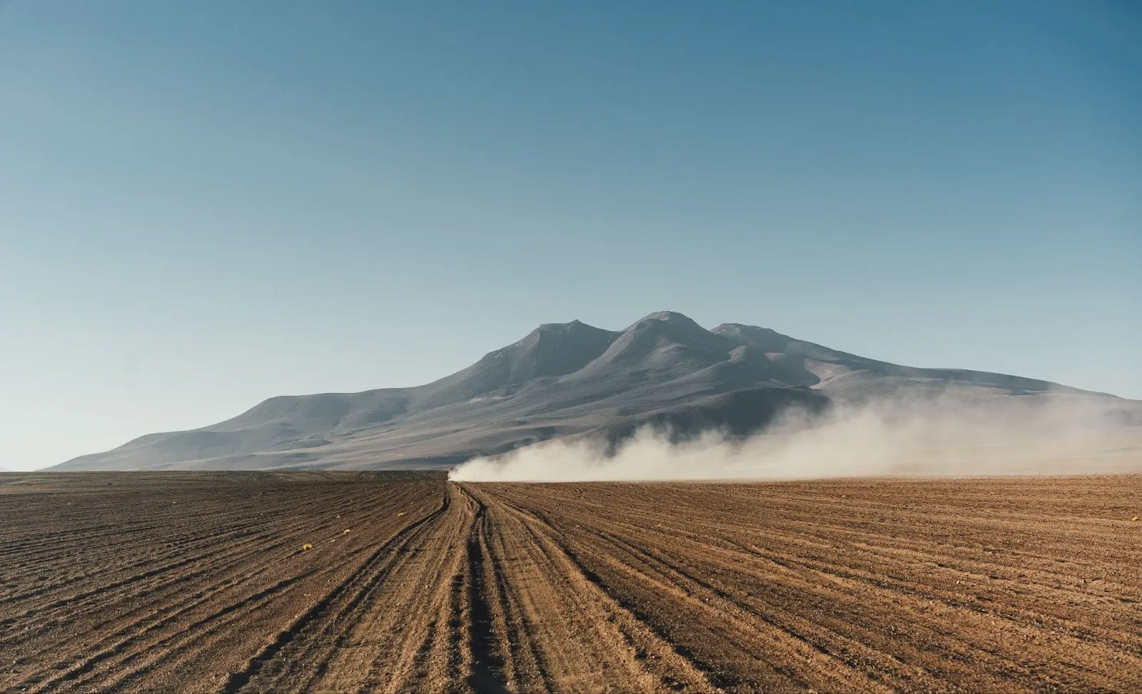 Dusty vehicle trail on a corrugated dirt road leading toward a volcanic mountain range under a clear sky.