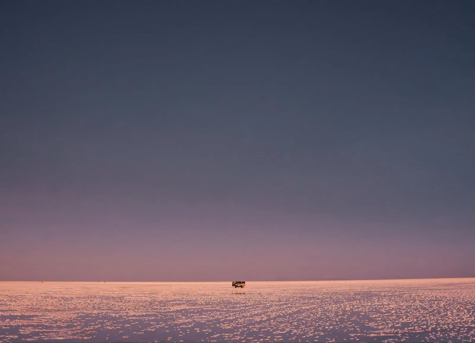 A distant off-road vehicle driving across a pink-tinted salt flat under a twilight sky.