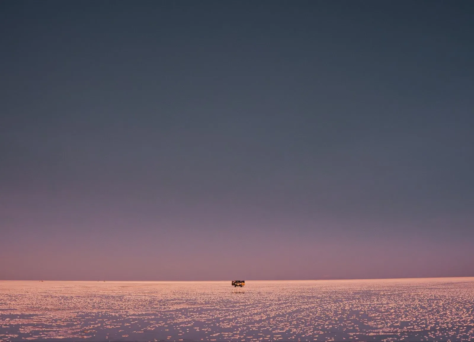 A distant off-road vehicle driving across a pink-tinted salt flat under a twilight sky.
