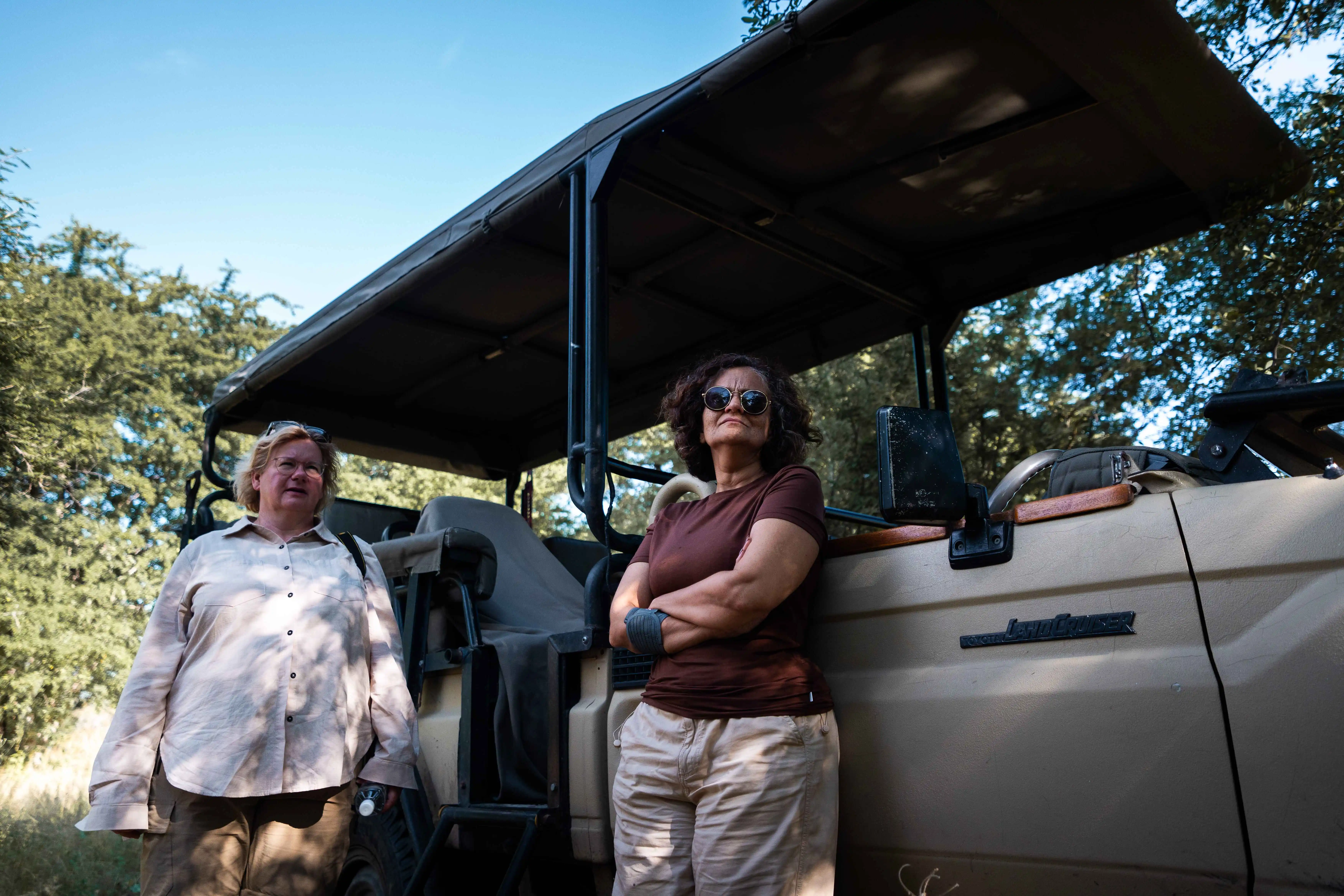 Two women travelers stand proudly beside an open-top 4x4 expedition vehicle under a canvas canopy during a Nomadic Road overland journey.