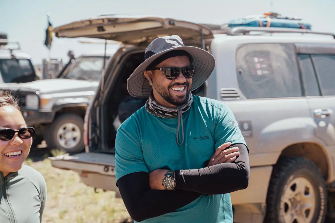 Nomadic Road expedition leader Swithun smiles during an overland journey, wearing a technical Columbia shirt, sun hat, and UV arm sleeves.
