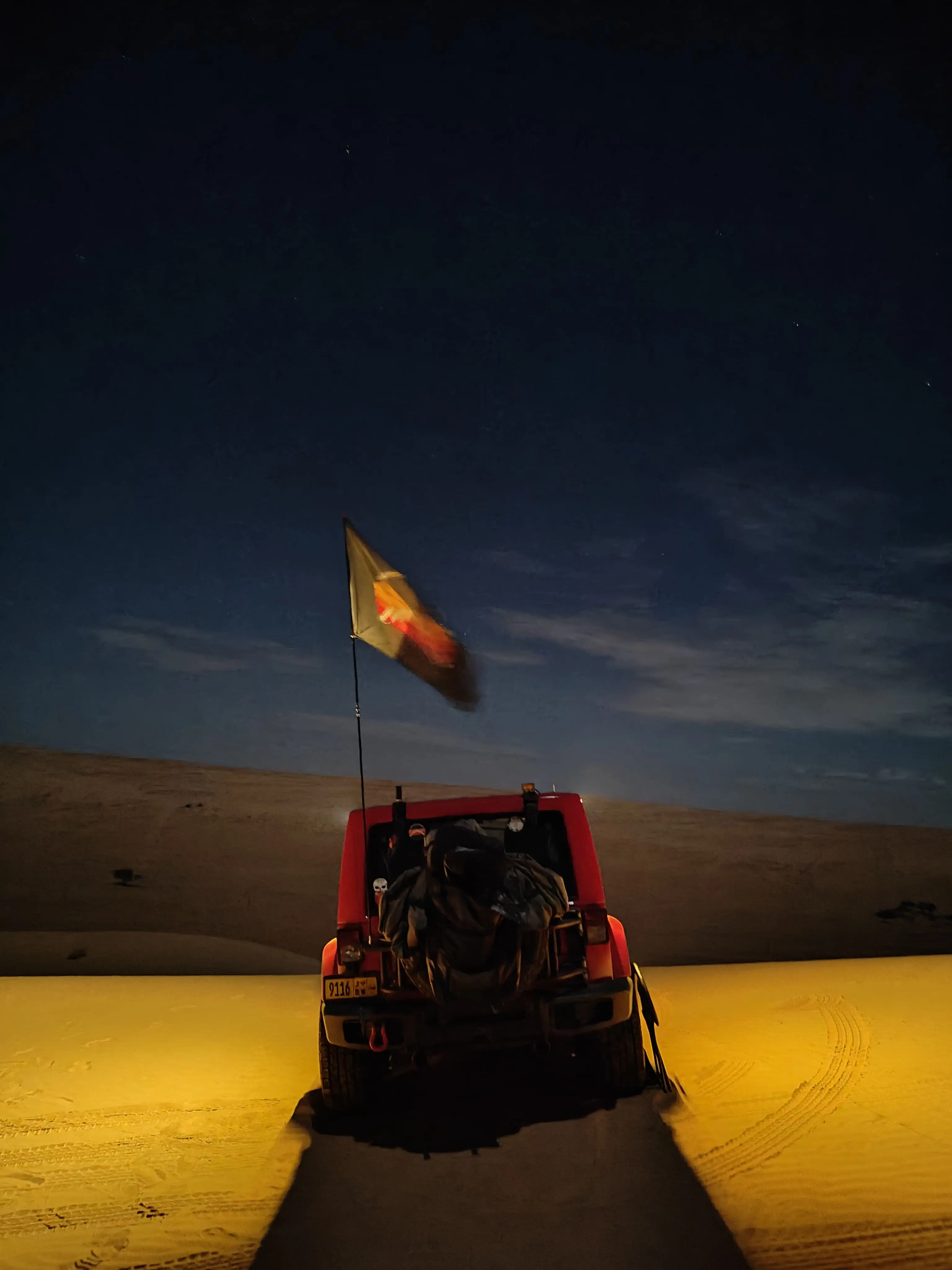 An off-road vehicle with a high-visibility safety whip flag attached to the rear bumper, parked on illuminated desert sand dunes under a dark night sky during a Nomadic Road expedition.