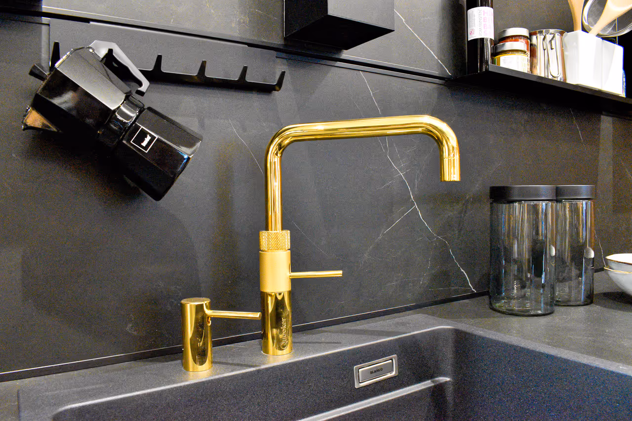 Close-up of a modern kitchen sink area with a gold tap, set against a black marble backsplash. A black coffee maker hangs on a rail above the sink, and glass jars are neatly placed on the counter.