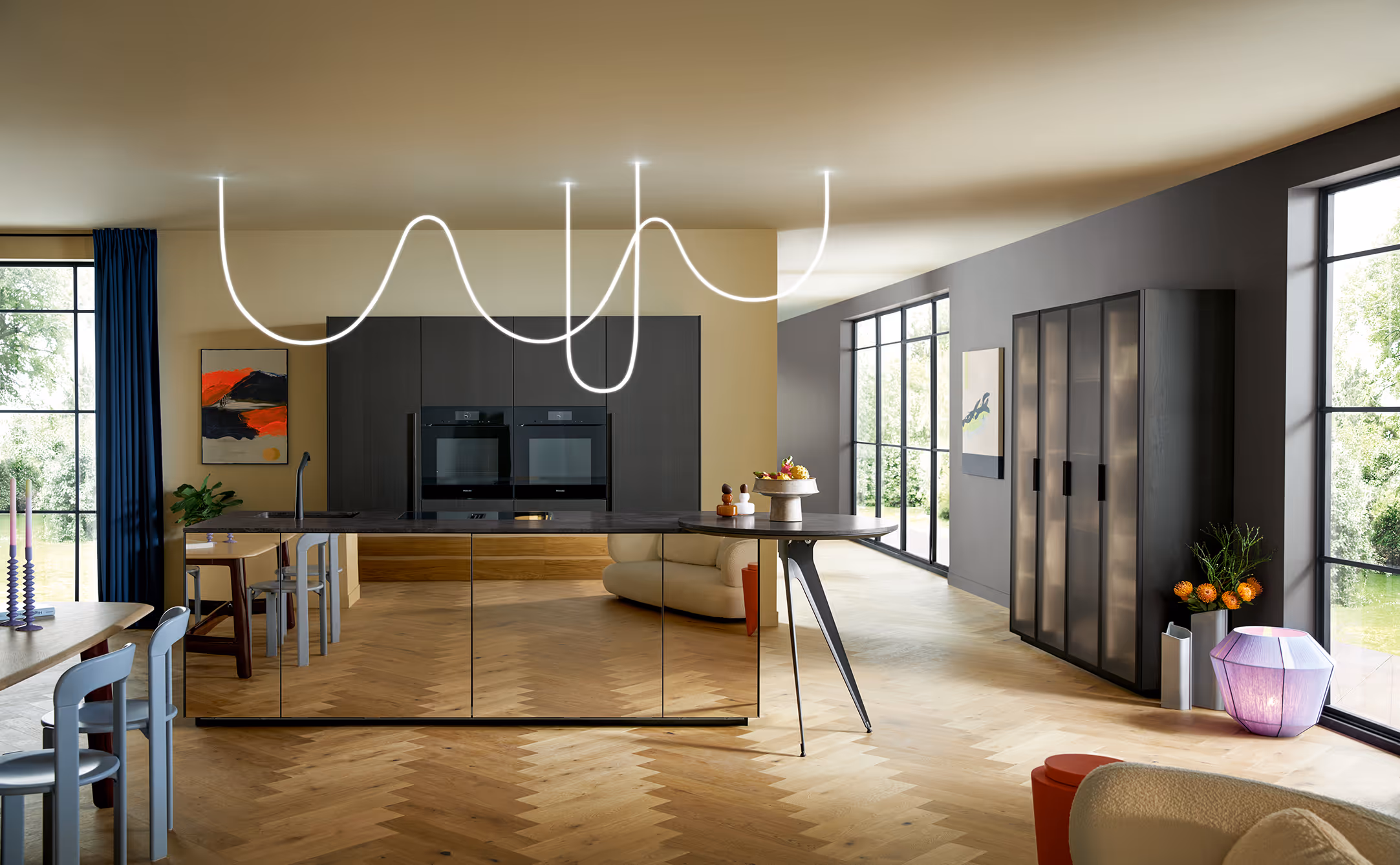 A spacious, modern kitchen with a sleek design, featuring a central island with reflective surfaces, integrated double ovens, and a unique ceiling light fixture. Large windows allow natural light to fill the space.

