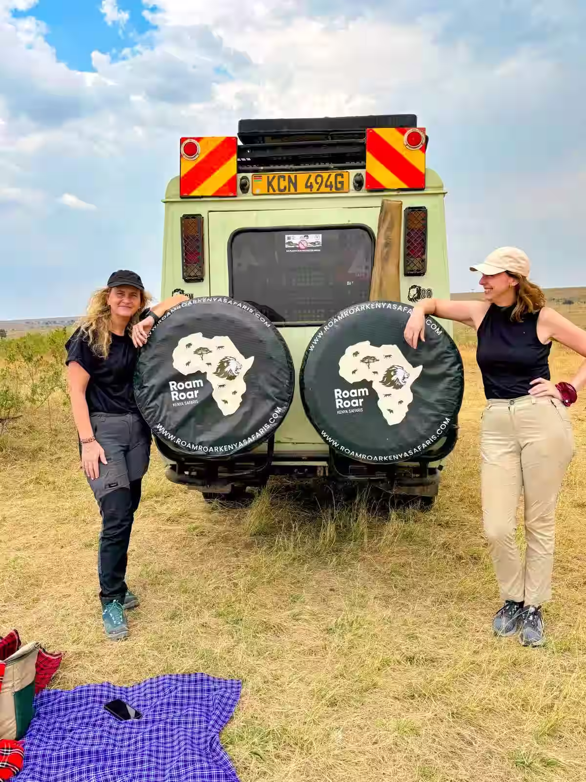 Two women standing on either side of a safari vehicle with spare tire covers showing a map of Africa and the text Roam Roar Kenya Safaris in a grassy savanna.