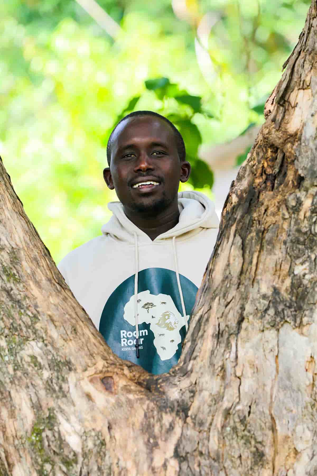 Smiling man wearing a roamroar hoodie standing between two tree trunks with green foliage in the background.