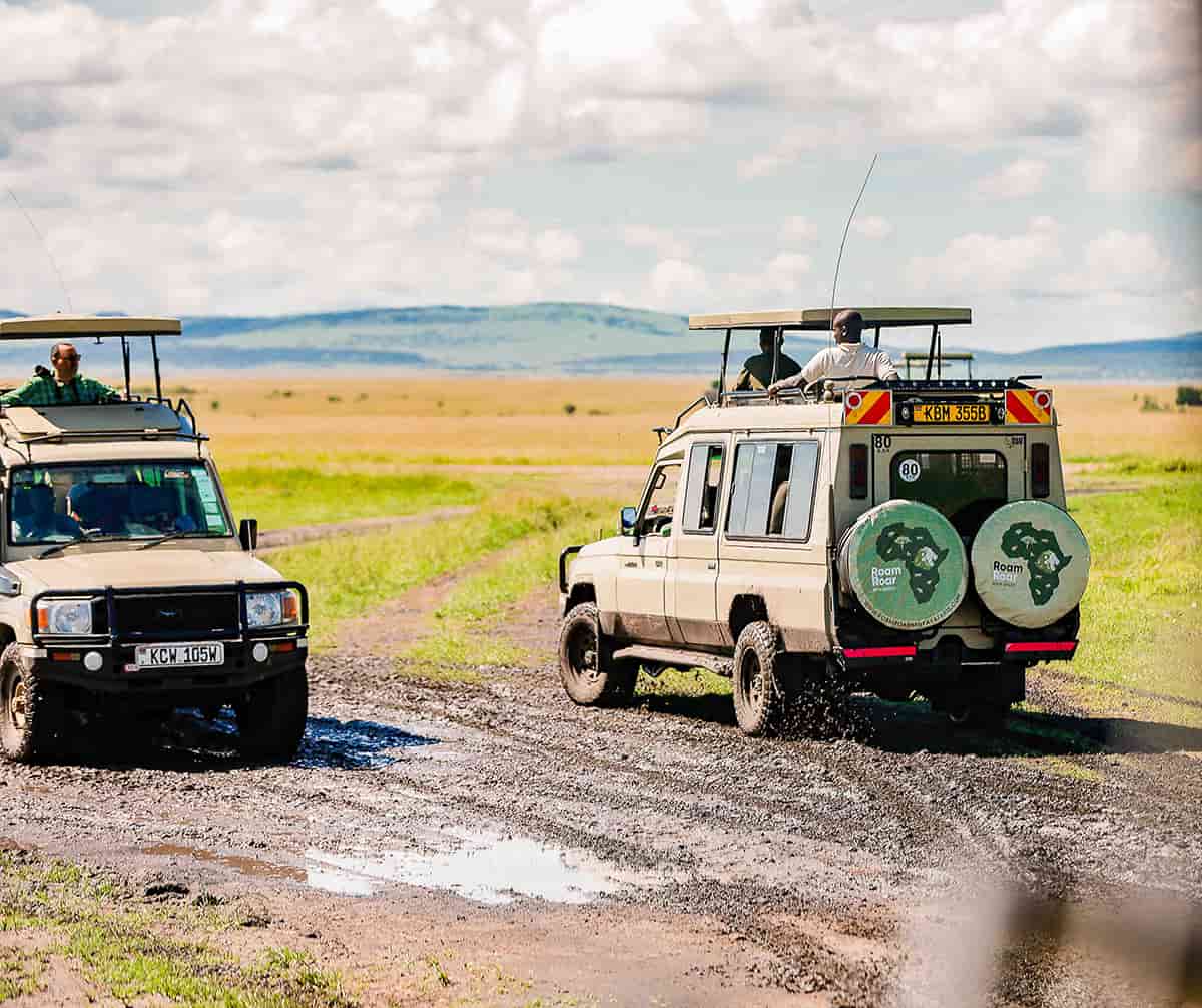 Two safari vehicles with open roofs driving on a muddy dirt road in a savanna landscape under a cloudy sky.